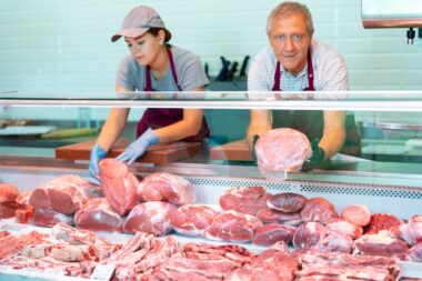 Smiling,experienced,aged,butcher,working,behind,counter,in,butchery,,showing